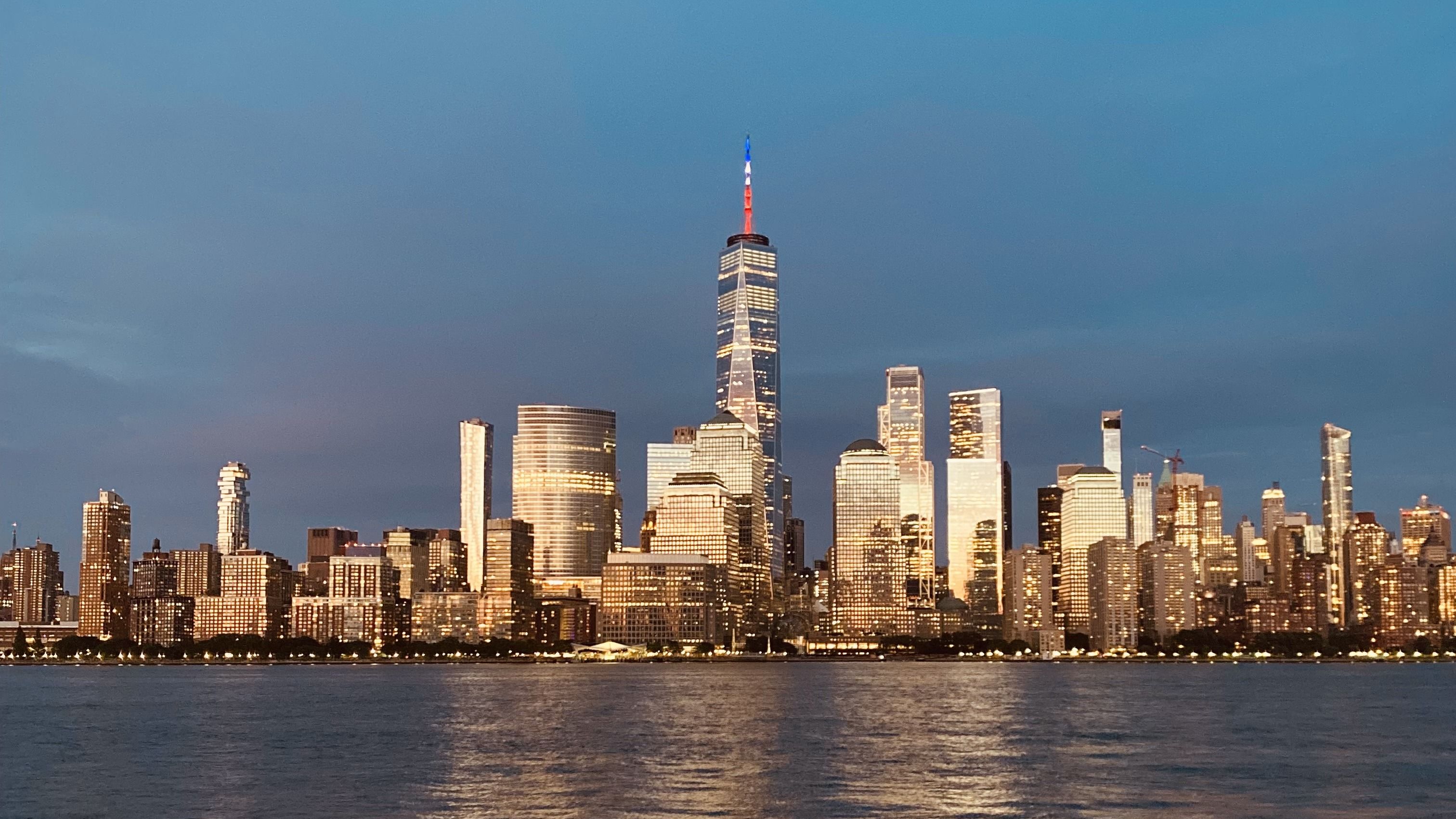 The Lower Manhattan skyline at dusk, lit windows reflected in the Hudson River